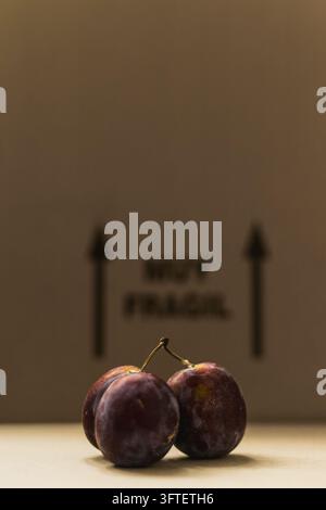 Two ripe dark purple plums on a cardboard surface with blurred fragile box marking and arrows in the background Stock Photo