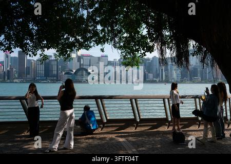People taking photos and relaxing on the Tsim Sha Tsui waterfront with Victoria Harbour and the Hong Kong Island skyline in the background, Hong Kong. Stock Photo