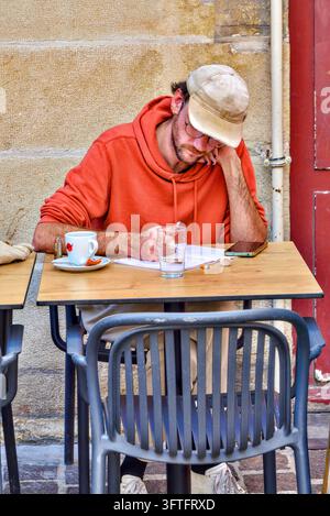 Young bearded man in red hoodie and baseball cap, leaning on elbow whilst writing notes - Tours, Indre-et-Loire (37), France. Stock Photo