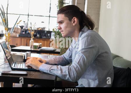 A young man sits at a cafe table focused on his laptop while sipping coffee. The scene takes place in a cozy cafe with plants and soft lighting Stock Photo