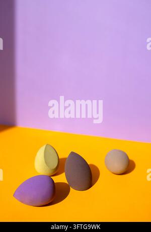 Four smooth Cosmetic sponges on yellow background, top view in yellow, purple, brown, and beige rest on a bright yellow surface before a lavender wall Stock Photo