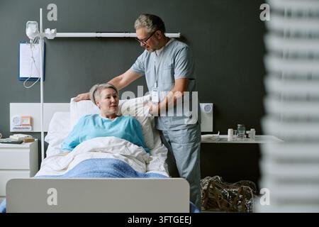 Male Nurse Adjusting Pillow for Mature Woman Patient in Hospital Bed Stock Photo