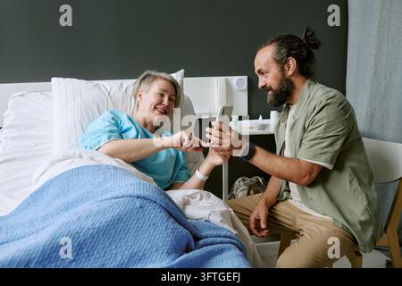 Middle Aged Woman Resting in Hospital Bed Looking at Smartphone with Man Stock Photo