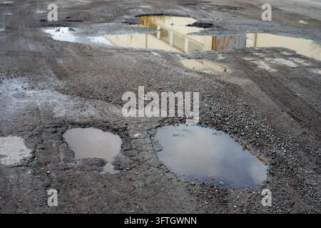 Potholes filled with rainwater on a damaged asphalt road. Cracked and deteriorating road surface in need of maintenance. Stock Photo