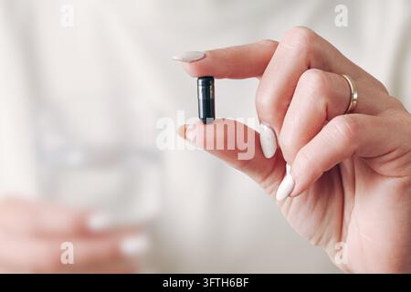 Woman holding a small black supplement capsule between fingers near a glass of water. Closeup of oral medication and daily healthcare routine. Stock Photo