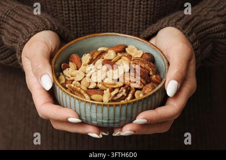 Hands holding a ceramic bowl filled with mixed nuts including cashews, almonds and pecans. Healthy snack assortment in a cozy home setting. Stock Photo
