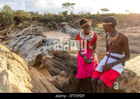 Smiling young Samburu tribe woman and man sitting and talking on rocks above a river gorge at sunset, Samburu County, Samburu, Kenya Stock Photo