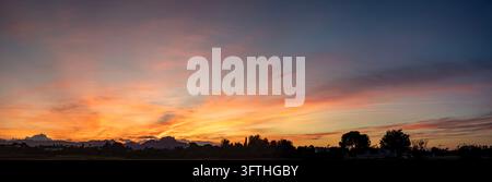 A wide panoramic landscape photograph capturing a dramatic sunset with vibrant orange, pink, and deep blue hues across a streaked cloudy sky. The hori Stock Photo