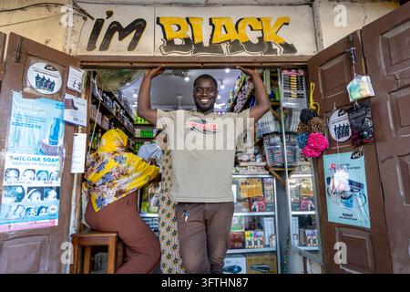 Smiling shopkeeper leaning on a cosmetics shop doorframe under the painted I am Black sign with a woman seated inside, Stone Town, Tanzania Stock Photo