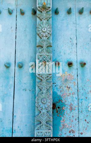 Close-up of a pale blue weathered wooden door with a carved floral strip and rusty studs and peeling paint in vertical view, Stone Town, Tanzania Stock Photo