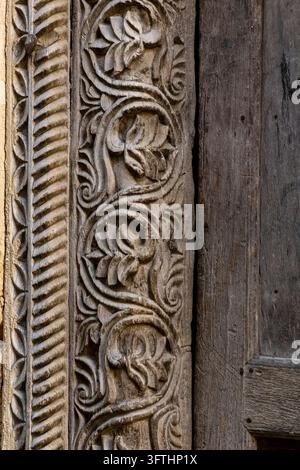 Close-up of intricate floral scroll carving on the wooden frame of an old door beside weathered grey planks, Stone Town, Tanzania Stock Photo