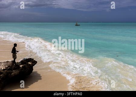 Silhouette of a young man in shorts standing on a coral rock above the surf looking at a traditional sailing dhow in stormy sky, Nungwi, Tanzania Stock Photo