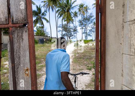 Man in a light blue polo shirt seen from behind pushing a bicycle through a rusty gate to a sandy path with palms and goats, Matemwe, Tanzania Stock Photo