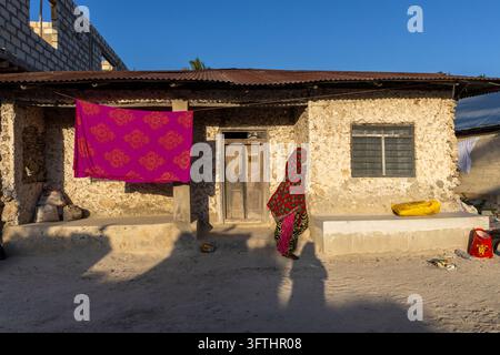 Woman in a red floral headscarf and long dress walking past a coral stone house with a wooden door and a pink cloth drying, Jambiani, Tanzania Stock Photo