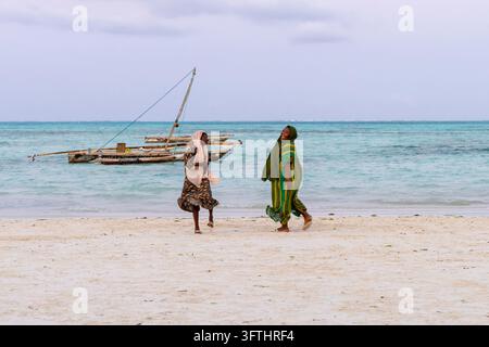 Two women in headscarves and long dresses laughing and running along a white sand beach with wooden dhows moored behind them, Jambiani, Tanzania Stock Photo