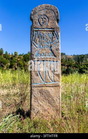 A close up view shows the carved face and blue painted inscriptions on a traditional stone memorial marker, Vakinankaratra, Antsirabe, Madagascar Stock Photo