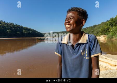 A smiling young man laughs beside the river as a boat sails past forested hills, Menabe, Tsiribihina, Madagascar Stock Photo