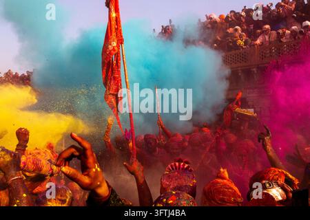 Clouds of blue yellow and pink powder over a crowd with a red flag at Braj Ki Holi, Uttar Pradesh, Nandgaon, India Stock Photo