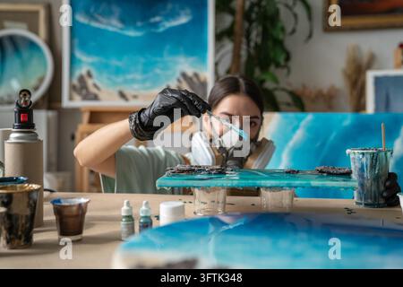 Woman crafter in respirator works with epoxy resin using canvas and spatula at table in studio, professional female artist adding liquid resin on painting, creating fluid art masterpiece in workshop. Stock Photo