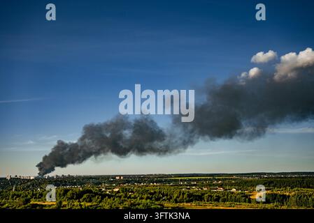 Thick black smoke billows into the clear blue sky, originating from a large fire burning within a residential or industrial area, creating an urban disaster and polluting the environment Stock Photo