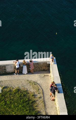 Top-down view of tourists standing on a stone lookout wall overlooking deep blue sea in Piran, Slovenia, near St. George's Church. Stock Photo