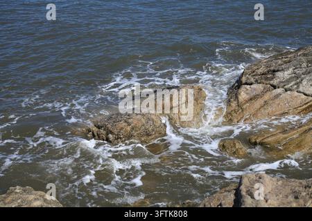 Sea water flows over rough coastal rocks, with white waves surging around them, showing peaceful yet dynamic natural coastal details. Stock Photo