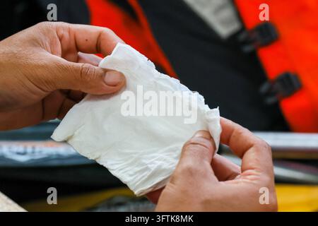 An adult hand holding white tissue paper during outdoor cleanup, with red safety jacket and equipment in background, showing practical hygiene and mai Stock Photo
