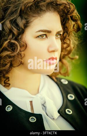 Side-profile portrait of a curly-haired woman in a black jacket over a white blouse, looking to the right against a softly blurred green background. Stock Photo