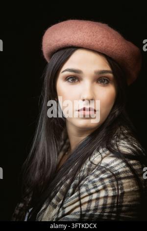A young woman wearing a brown beret and plaid coat looks directly toward the camera in a dramatic studio portrait against a black background. Stock Photo