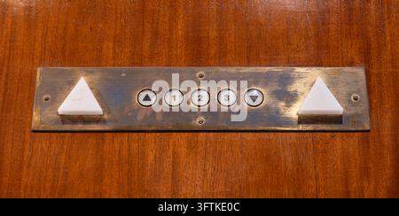 Close-up of a vintage elevator control panel featuring numbered floor buttons, directional arrows, and a worn metal plate mounted on a wooden surface. Stock Photo