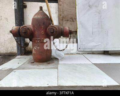 Weathered rust-covered fire hydrant stands on cracked tiled pavement beside a stained concrete wall and utility panel Stock Photo