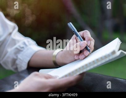 Asian man hands with pen writing on notebook in park. Writing notebook diary, working from anywhere concept. Stock Photo