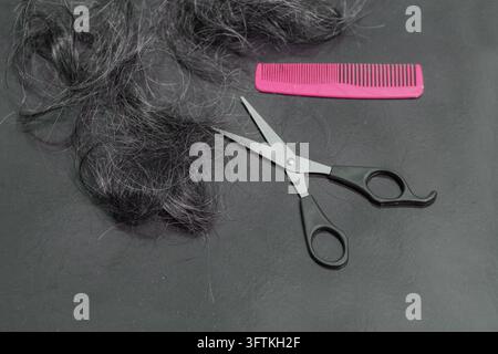 Hairdresser's scissors with pink comb and strand of blonde hair on black table background. Top view. Stock Photo