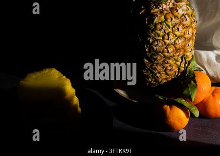 Pineapple on a rustic wooden log with mandarin oranges stacked around it next to sliced ​​pineapple on a black plate on a dark background. Dark fruit Stock Photo
