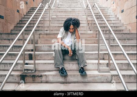 Young Black man sitting on concrete stairs with head in hand, looking down, feeling sad Stock Photo