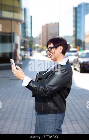 A smiling mature businesswoman in profile walks along a modern Helsinki street, using her smartphone and holding a laptop. Stock Photo