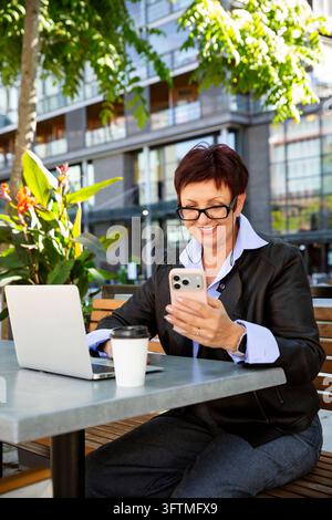 A smiling mature businesswoman uses a phone and laptop at an outdoor cafe in Helsinki, a senior digital nomad enjoying remote work. Stock Photo