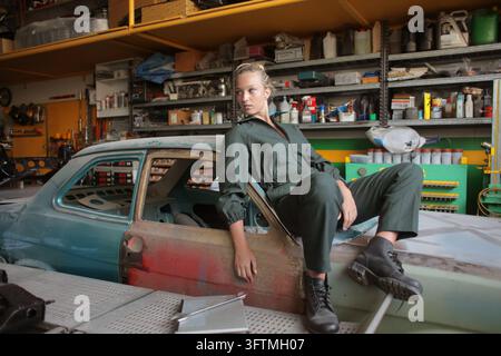 Confident young woman in a jumpsuit sits on a vintage car Stock Photo