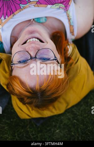 Happy woman with red hair and glasses lying on grass Stock Photo