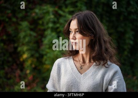 Young woman in white sweater looking away against green leaves Stock Photo