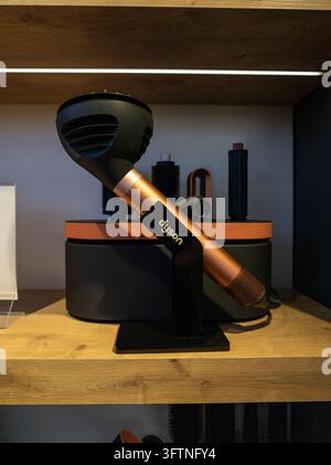 Black and copper hair dryer on wooden shelf. Dryer stands upright on black stand. Background shows other hair styling tools. Shelf wood grain texture Stock Photo