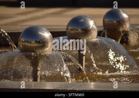 Close-up view of reflective spherical ornaments above flowing water in  outdoor fountain. Warm sunlight highlights the splashing streams, wet surface Stock Photo