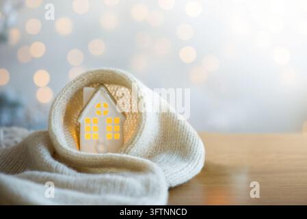 Miniature house with glowing windows wrapped in a warm knitted blanket on a wooden table. Cozy winter home and heating concept with copy space. Stock Photo