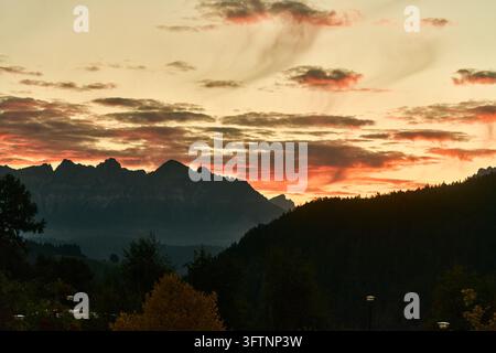 Dolomites mountain ridge silhouette over forested hills at dramatic orange and gold sunset with layered clouds and foreground trees and lamps Stock Photo
