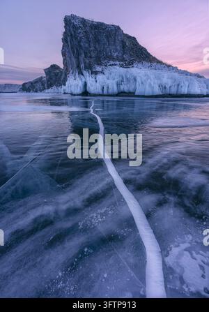 View of Lake Baikal with frozen ice, large cracks in the ice, and rocky cliffs under a soft pink sunset sky, Russia. Stock Photo