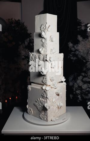 Four-tier square wedding cake covered in smooth white fondant with cascading edible white sugar flowers and delicate floral relief decorations, displa Stock Photo