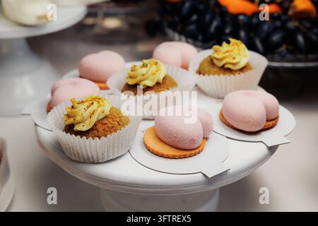 Close-up of assorted pastries displayed on white cake stand including pistachio cupcakes with piped yellow frosting in paper liners and pink sugar-coa Stock Photo