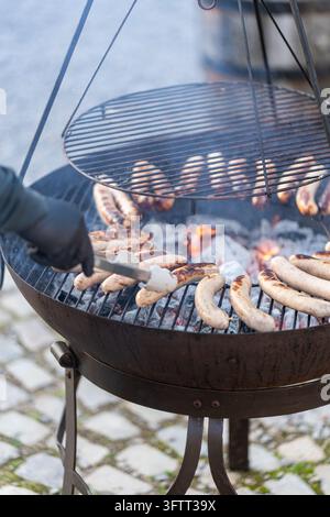 Sausages grilling over glowing charcoal on a fire pit barbecue with hanging grill, turned with tongs by a gloved hand on cobblestones Stock Photo