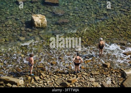 Three women standing on a rocky beach by crystal clear waters of the Adriatic Sea in Piran, Slovenia. Stock Photo