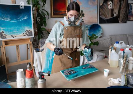Female artist holds plastic cup filled with blue epoxy resin and carefully drips liquid onto painting with wooden spatula, controlling placement to create desired pattern in home loft workshop. Stock Photo